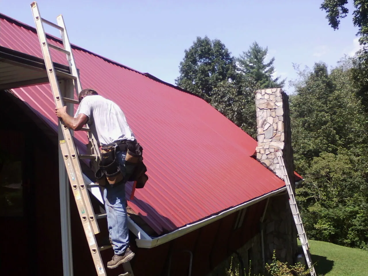 Roofer on a ladder beside a red metal roof and stone chimney — Soffit & Fascia Repair by Roofing Brothers in Jacks Creek